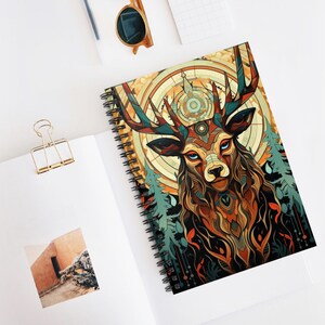 May include: Spiral-bound notebook featuring a colorful illustration of a stag with intricate patterns and blue eyes. The design incorporates a compass and forest elements, with a palette of orange, teal, and brown. The notebook is open on a white surface.