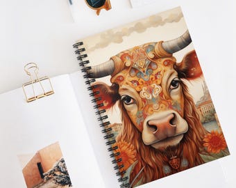 Könnte beinhalten: Spiralgebundenes Notizbuch mit einer detaillierten Illustration einer Kuh mit kunstvollen, farbenfrohen Mustern auf Kopf und Körper. Die Kuh hat große Hörner und ist von floralen Elementen umgeben. Das Notizbuch liegt offen auf einer weißen Oberfläche.