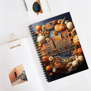 May include: Spiral-bound notebook with a vibrant illustration of a Dutch village, including miniature buildings and food items, set against a dark blue backdrop. The notebook is open on a white surface, with a gold paper clip and sunglasses visible.