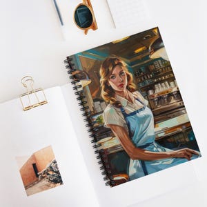 Op de afbeelding: Spiraalgebonden notitieboek met een retro-illustratie van een restaurant. De afbeelding toont een vrouw in een wit overhemd en een blauw schort. Het notitieboek ligt open op een wit oppervlak met een gouden paperclip en zonnebril.