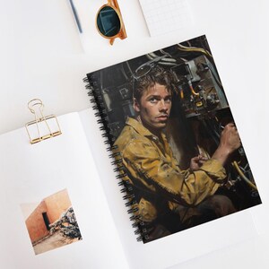 May include: Spiral-bound notebook featuring a detailed illustration of a person in a yellow work uniform, operating machinery. The image has a vintage aesthetic. The notebook is open on a white surface, with a gold paper clip and sunglasses nearby.