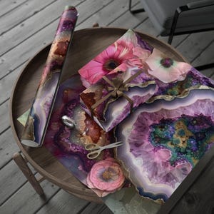 May include: A roll of patterned wrapping paper and wrapped gifts with a geode design in shades of purple, pink, and gold. A pair of gold scissors and a silver object are also on the wooden table.