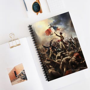 May include: Spiral-bound notebook with a historical painting on the cover, depicting a battle scene with figures holding a flag. The artwork shows a dramatic scene with a woman holding a flag, and soldiers in the background. The notebook is open on a white surface.