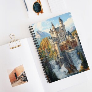 Può includere: Quaderno a spirale con un'illustrazione dettagliata di un castello su una scogliera con cascate. L'opera d'arte raffigura un maestoso castello con più torri, circondato da montagne e fogliame autunnale. Il quaderno è aperto su una superficie bianca.