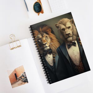 Puede incluir: Cuaderno de espiral con una ilustración de dos leones con ropa formal. Uno tiene una melena dorada, el otro una melena blanca. Ambos llevan esmóquines y pajaritas. El cuaderno está abierto sobre una superficie blanca.