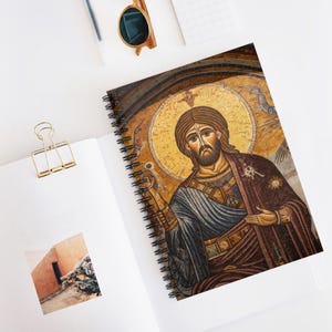 Könnte beinhalten: Spiralgebundenes Notizbuch mit einem Mosaikbild einer religiösen Figur. Das Kunstwerk zeigt braune, blaue und goldene Farbtöne. Das Notizbuch liegt offen auf einer weißen Oberfläche, mit einer goldenen Büroklammer und Sonnenbrille.