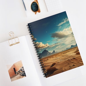 May include: Spiral-bound notebook with a desert landscape design, featuring a blue sky, clouds, and mountains. The notebook is open, showing a white page and a gold paper clip. Sunglasses are in the background.