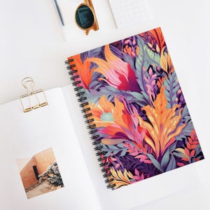 May include: Spiral-bound notebook with a vibrant floral design. The cover features a colorful illustration of stylized flowers and foliage in shades of orange, pink, purple, and green. The notebook is open, revealing blank pages and a photo.