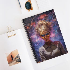 May include: Spiral-bound notebook with a digital art portrait of a woman with blonde hair and sunglasses. The artwork has a vibrant, colourful background with a futuristic theme. The notebook is open on a white surface.
