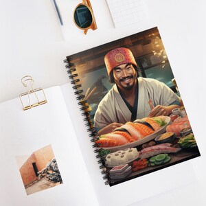Könnte beinhalten: Spiralgebundenes Notizbuch mit einer Illustration eines lächelnden Sushi-Kochs. Der Koch, in weißer Uniform und roter Mütze, ist von Sushi umgeben. Das Notizbuch liegt offen auf einer weißen Oberfläche, mit einer goldenen Büroklammer und Sonnenbrille.
