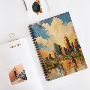 May include: Spiral-bound notebook featuring an impressionistic painting of a cityscape with tall buildings, a reflective body of water, and a colorful sky. The notebook is open on a white surface, with a gold paper clip and sunglasses nearby.