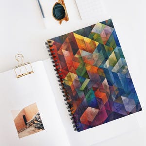Może przedstawiać: Spiralny notatnik z kolorowym geometrycznym wzorem. Okładka przedstawia mozaikę kanciastych kształtów w odcieniach czerwieni, pomarańczy, żółci, zieleni, niebieskiego i fioletu. Notatnik jest otwarty, ukazując puste białe strony.