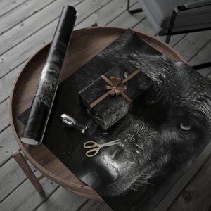 Op de afbeelding: Een ronde houten tafel met een rol zwart-wit cadeaupapier, een ingepakt cadeau met een bruin lint, een schaar en een tape dispenser. Het cadeaupapier en het cadeau tonen een close-up van het gezicht van een wolf.