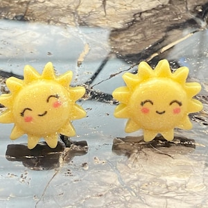 May include: Two yellow glitter sun shaped resin earrings with a smiling face and pink cheeks.