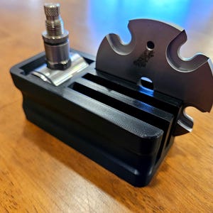 May include: A black 3D-printed razor stand with a stainless steel razor and blade. The stand has slots for the razor and blade. The blade is a unique shape with a decorative design. The stand is on a wooden surface.