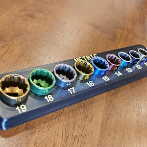 May include: A black socket organizer with a variety of colored sockets. The sockets are arranged in a row and labeled with metric sizes from 10 to 19. The sockets are gold, green, blue, red, and silver.