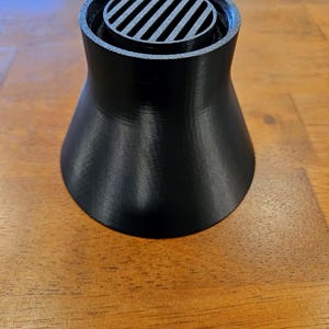 May include: A black, 3D-printed funnel-shaped object with a slatted top. The base is wider than the top, creating a flared design. The object is sitting on a wooden surface.