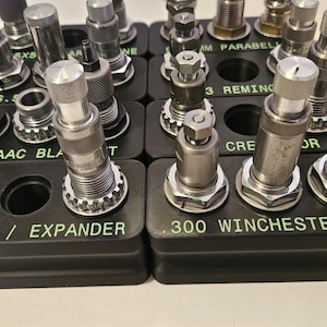 May include: A collection of reloading tools, including expanders and dies, arranged in black holders. The tools are made of silver metal and are labeled with text such as ".300 AAC BLACKOUT" and ".300 WINCHESTER MAG". The text "REPRIME / EXPANDER" is also visible.