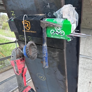 Puede incluir: Un conjunto de herramientas de construcci&oacute;n, que incluye una amoladora angular roja, un cepillo de alambre, un martillo con mango azul y una caja de varillas de soldadura. La caja es verde y tiene el texto "5/32 4.0mm x 400mm". Tambi&eacute;n se ve una caja negra con el texto "RECONSTRUCT".