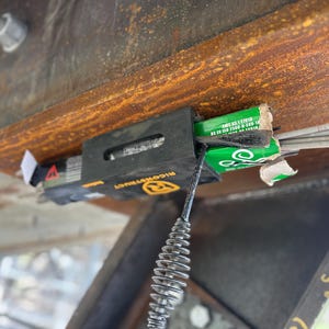 Puede incluir: Vista de cerca de un portaelectrodos negro y amarillo sujeto a una viga met&aacute;lica oxidada. El soporte contiene cajas verdes y blancas de electrodos y una bobina met&aacute;lica. La viga tiene un perno y una tuerca.