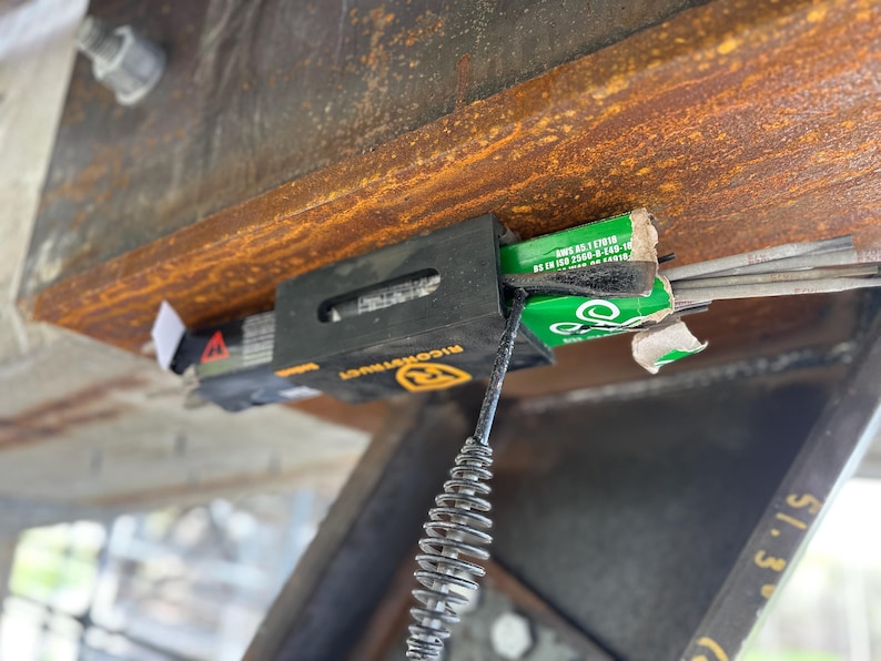Puede incluir: Vista de cerca de un portaelectrodos negro y amarillo sujeto a una viga met&aacute;lica oxidada. El soporte contiene cajas verdes y blancas de electrodos y una bobina met&aacute;lica. La viga tiene un perno y una tuerca.