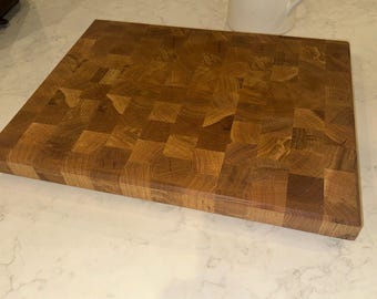 May include: A large, square end-grain chopping board. The board is made of various pieces of wood, creating a checkerboard pattern. The wood has a warm, brown tone. A white jug is in the background.