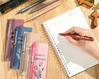 May include: A collection of pens and notebooks on a wooden surface. Pens in various colors, including pink, gray, and brown, with inspirational quotes. A hand holds a red pen, writing on a spiral-bound notebook. Decorative wooden crosses and books are also present.
