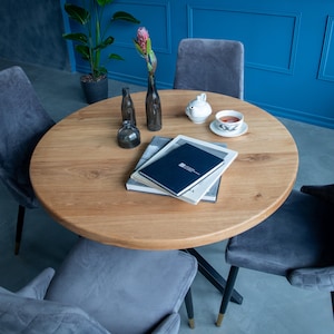 Puede incluir: Una mesa redonda de madera clara, rodeada de sillas de terciopelo gris. La mesa está decorada con libros, una taza de té, una tetera y botellas de vidrio decorativas. Un jarrón con una flor rosa también está sobre la mesa.