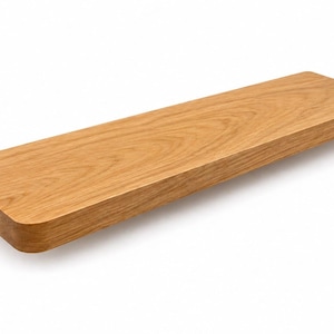 May include: A rectangular wooden shelf with rounded corners. The shelf is made of light brown wood and has a natural wood grain pattern. The shelf is designed to be mounted on a wall and is suitable for displaying items.