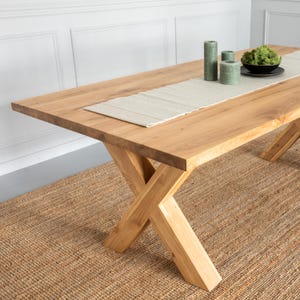 Op de afbeelding: Een grote, rechthoekige houten eettafel met een natuurlijke afwerking. De tafel heeft een kruisvormige poot en een lichtgekleurde loper. Groene kaarsen en een kleine kom met groen zijn op de tafel geplaatst.