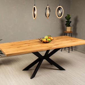 May include: A wooden dining table with a black metal X-shaped base. The table top is made of light wood and has a natural finish. The table is set with a bowl of fruit.