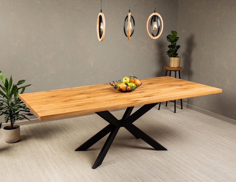 May include: A wooden dining table with a black metal X-shaped base. The table top is made of light wood and has a natural finish. The table is set with a bowl of fruit.