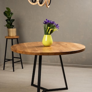 Op de afbeelding: Een ronde houten tafel met een zwart metalen onderstel. De tafel is gedekt met een vaas met paarse bloemen en een gele vaas.