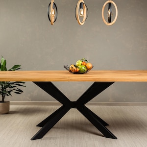 May include: A large wooden dining table with a black metal X-shaped base. The table is set with a bowl of fruit and has a light wood finish.