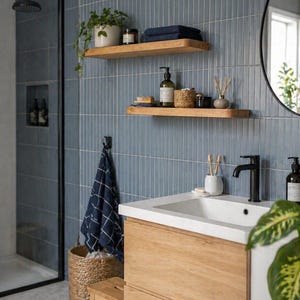 May include: A modern bathroom with blue tile walls, wooden shelves, and a wooden vanity. The shelves hold plants, towels, and toiletries. A black faucet and a round mirror are also visible. A woven basket and a small wooden stool add to the decor.