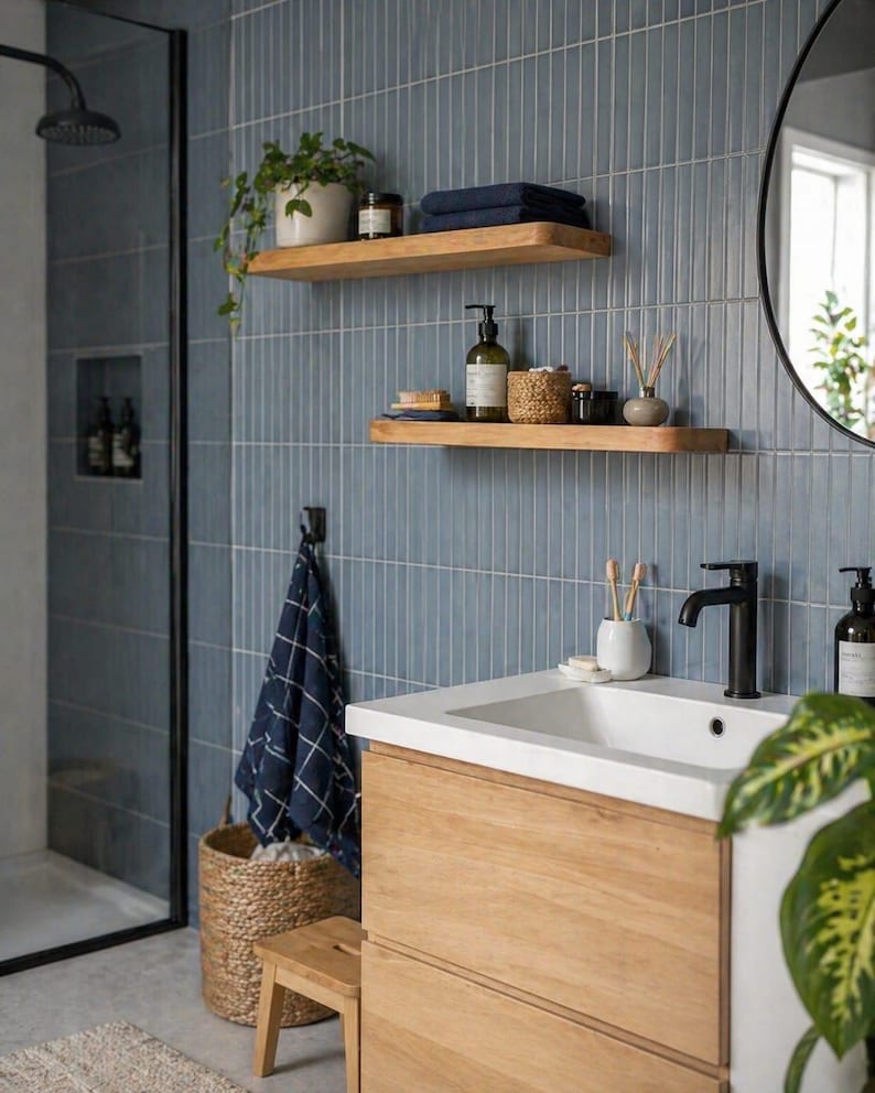May include: A modern bathroom with blue tile walls, wooden shelves, and a wooden vanity. The shelves hold plants, towels, and toiletries. A black faucet and a round mirror are also visible. A woven basket and a small wooden stool add to the decor.