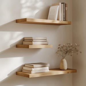 Puede incluir: Tres estantes flotantes de madera montados en una pared blanca. Los estantes son de madera clara y contienen pilas de libros y un jarr&oacute;n con flores secas. Los estantes est&aacute;n dispuestos en una esquina.