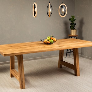 May include: A large, rectangular wooden dining table with a natural finish. The table has a sturdy, trestle-style base. A bowl of fruit sits on the table's surface. Overhead, three pendant lights hang from the ceiling, illuminating the space.