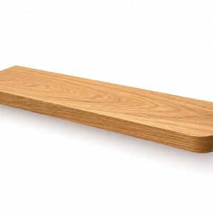 May include: A rectangular wooden shelf with rounded corners. The shelf is made of light-colored wood, showcasing a natural grain pattern. It is designed to be mounted on a wall, providing a simple and functional storage solution. The shelf is approximately 30 inches long.