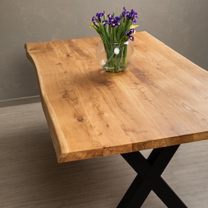 Op de afbeelding: Een houten eettafel met een live edge blad en zwarte metalen X-vormige poten. Een glazen vaas met paarse bloemen staat op de tafel.