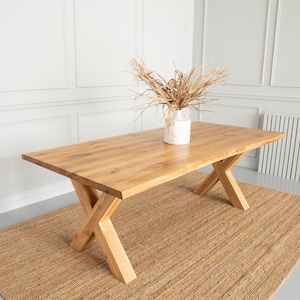 Könnte beinhalten: Ein rechteckiger Esstisch aus Holz mit natürlicher Oberfläche und kreuzförmigen Beinen. Eine weiße Vase mit getrockneten Pflanzen steht auf dem Tisch. Der Tisch steht auf einem gewebten Jute-Teppich.