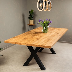May include: A wooden dining table with a natural finish and black X-shaped legs. The table is set with a vase of purple flowers.