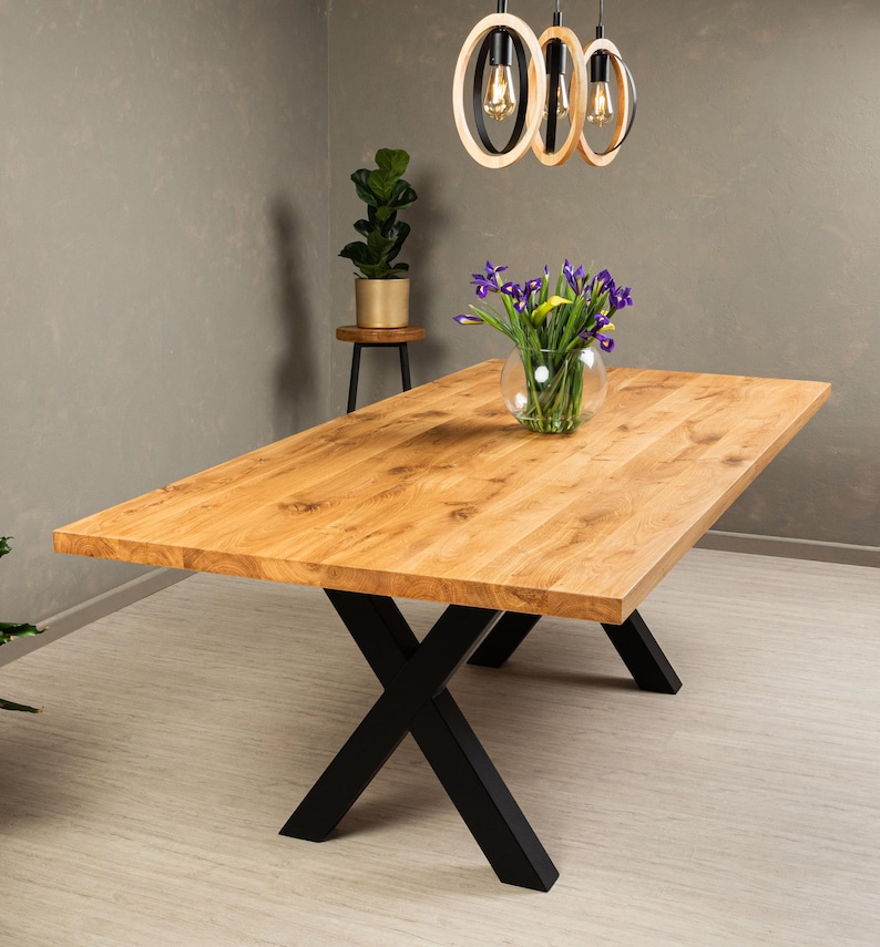 May include: A wooden dining table with a natural finish and black X-shaped legs. The table is set with a vase of purple flowers.