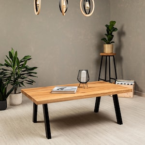 May include: A wooden coffee table with black metal legs. The table has a natural wood finish and is sitting on a light wood floor. There is a book and a decorative candle holder on the table.