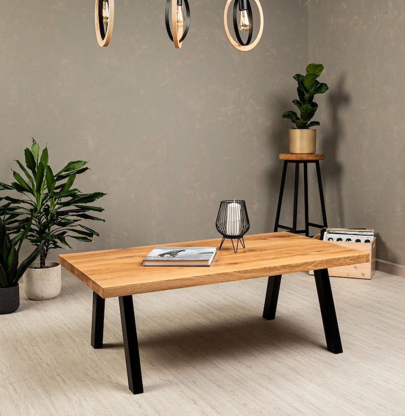 May include: A wooden coffee table with black metal legs. The table has a natural wood finish and is sitting on a light wood floor. There is a book and a decorative candle holder on the table.