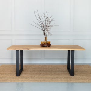 Könnte beinhalten: Rechteckiger Esstisch aus hellem Holz mit schwarzen Metallbeinen. Auf dem Tisch steht eine Vase mit Zweigen. Der Tisch steht auf einem gewebten Teppich. Die Tischlänge beträgt ca. 180 cm.