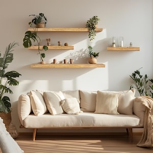 Puede incluir: Un sof&aacute; beige claro con varios cojines color crema y un marco de madera. Encima del sof&aacute;, tres estantes de madera exhiben plantas en macetas y objetos decorativos. La habitaci&oacute;n es luminosa y espaciosa, con luz natural.