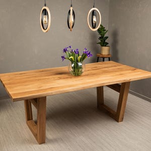 May include: A large, rectangular wooden dining table with a natural finish. The table has a sturdy, angled leg design. A glass vase of purple flowers sits on the table. Overhead, three pendant lights with circular wooden frames hang from the ceiling.