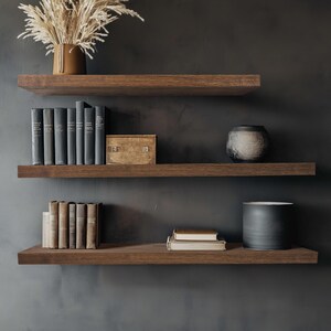 Puede incluir: Tres estantes flotantes de madera oscura contra una pared gris oscuro. Los estantes exhiben libros, jarrones decorativos, una caja peque&ntilde;a y una maceta de cer&aacute;mica con plantas secas. Los estantes son de color marr&oacute;n intenso, creando una est&eacute;tica moderna y minimalista.