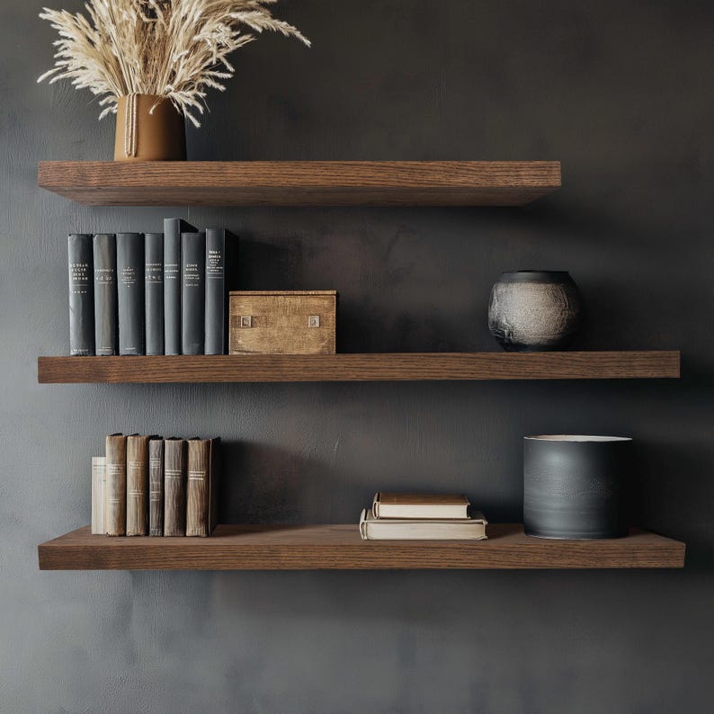 Puede incluir: Tres estantes flotantes de madera oscura contra una pared gris oscuro. Los estantes exhiben libros, jarrones decorativos, una caja peque&ntilde;a y una maceta de cer&aacute;mica con plantas secas. Los estantes son de color marr&oacute;n intenso, creando una est&eacute;tica moderna y minimalista.